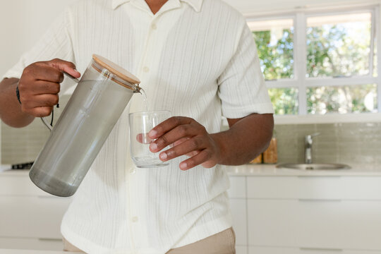 African American male wearing cream shirt pouring water from metal pitcher into glass kitchen sink