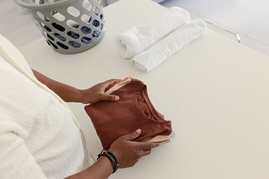 Man wearing light short-sleeve shirt folding rust-colored T-shirt on pale folding table, copy space