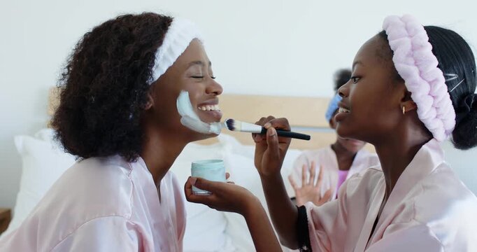 Younger female dipping brush into jar, applying facial mask, spreading down jawline for skincare