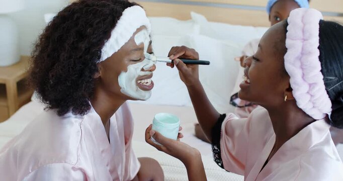 Vertical video: Dipping brush, pink-robed woman applying mask to recipient on bed bonding with jar