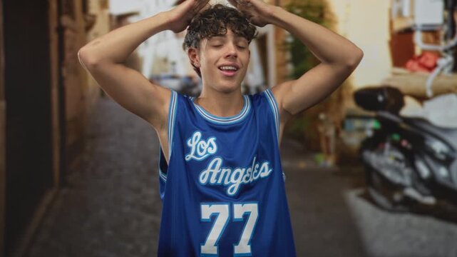 Man in blue los angeles basketball jersey with arms raised above head on a street showing bare shoulders and number 77; confidence youth sporty.