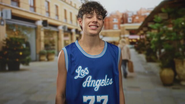 Teen boy in los angeles basketball jersey showing three fingers with hand raised on a street lined with shops and potted plants; playful confidence.