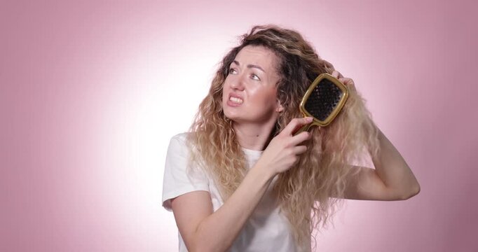 Unhappy woman trying to brush her tangled hair on pink background