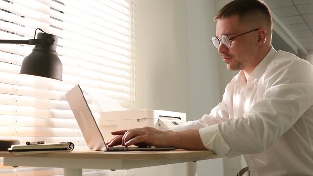 Man using printer while working on laptop at wooden desk indoors
