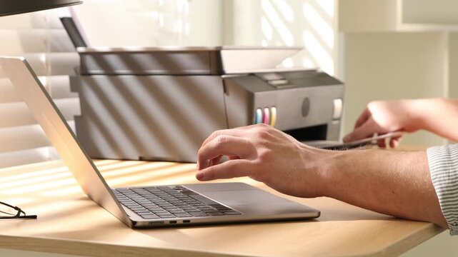 Man using printer while working on laptop at wooden desk indoors, closeup