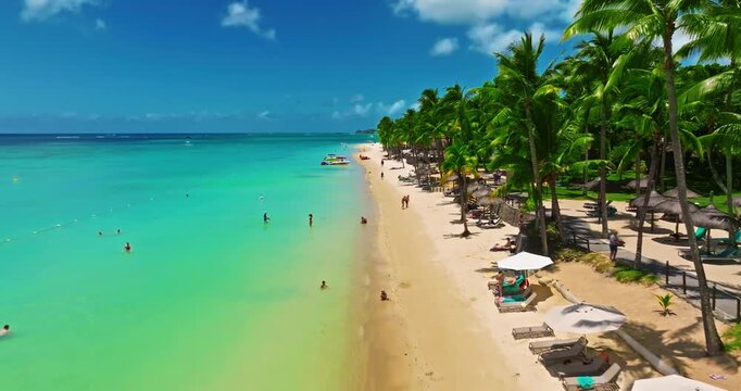 Aerial view of crystal water on the seashore, Mauritius with palm trees and living reef around the island. Establishing shot of sea lagoon formed by coral reefs. Tourists stroll along a tropical beach