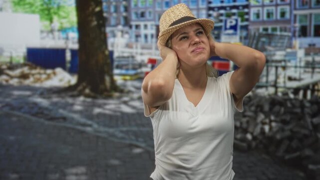 Woman covers ears with hands on street wearing straw hat and white t shirt by tree and parked bikes; annoyance stress.