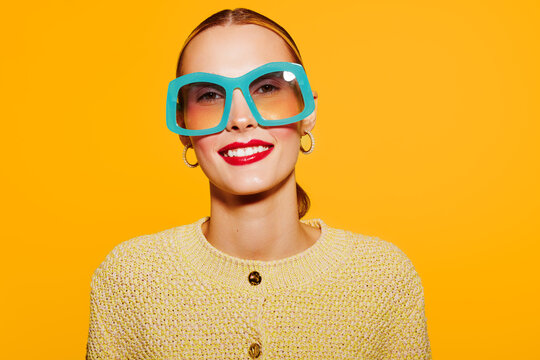 Smiling woman with large blue sunglasses and red lipstick wears yellow knitted sweater against bright yellow background. Concept of fashion, style, beauty, and vibrant colors.
