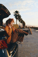 Naklejka premium Young woman with curly hair smiling and sitting in the open trunk of a car wearing a rainbow sweater, enjoying a relaxed lifestyle outdoors during golden hour near palm trees.