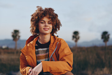 Smiling woman with curly hair wearing a rainbow sweater and orange jacket sitting outdoors near...