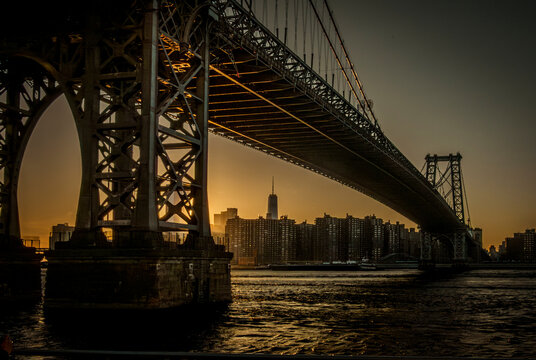 A stunning sunset view under the massive steel structure of the Williamsburg Bridge in NYC, with the Manhattan skyline and One World Trade Center visible in the hazy distance.&nbsp;