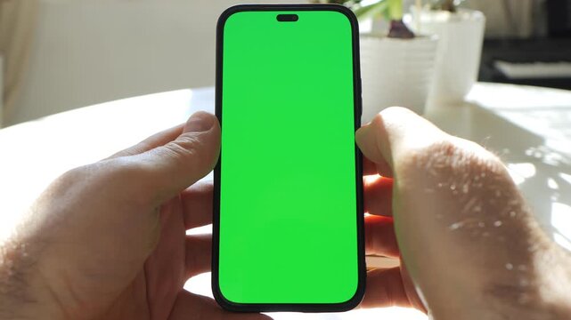 Hands hold phone with green screen. Fingers tap screen. Sunlight reflects off phone. Background shows blurred indoor plants. No animals or plants are present