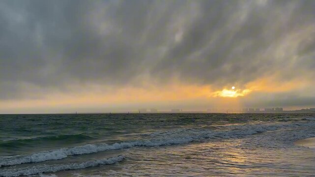 High frame rate 4K video of rhythmic waves on a beach at dusk. The sun glows through misty clouds over the modern skyline of a coastal city in Hainan, China.