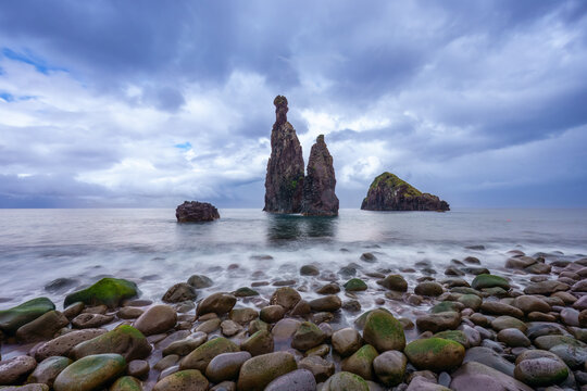 Long exposure of the dramatic Ilheus da Ribeira da Janela sea stacks and mossy volcanic rocks on a pebble beach under a moody sky, North Coast of Madeira Island, Portugal.