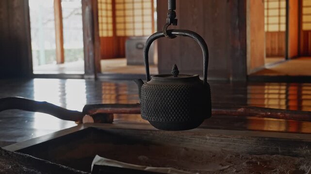 A detailed shot of a black cast iron kettle hanging over a traditional sunken hearth (irori). The background shows a blurry view of a classic Japanese house.