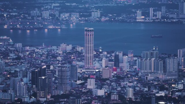 A high-angle day-to-night timelapse view showing the illuminated urban skyline of George Town, featuring the prominent Komtar Tower and city lights reflecting on the water.