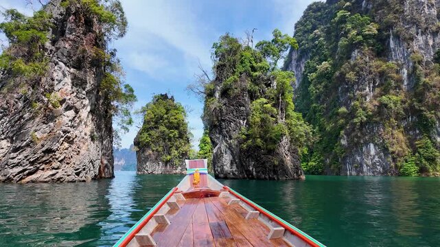 View from a traditional longtail boat approaching towering limestone karst pillars rising from emerald water on Cheow Lan Lake in Khao Sok National Park, Thailand.