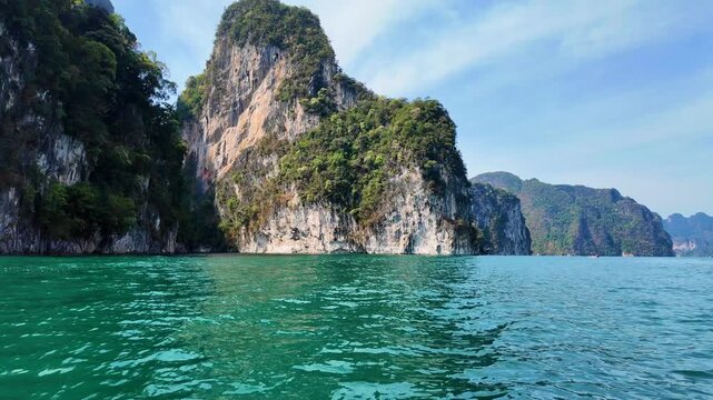 Massive limestone karst cliff rising above emerald green water surrounded by tropical rainforest on Cheow Lan Lake in Khao Sok National Park, Thailand.