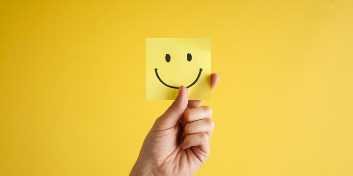 Hand holding yellow sticky note paper with happy smiling face on bright yellow background with copy space