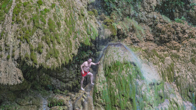 A picturesque waterfall. Thin streams of water flow down the mossy terraces, forming a veil. A man stands on a ledge under the drops, leans on his hand, looks at the camera. Philippines. Tumalog Falls
