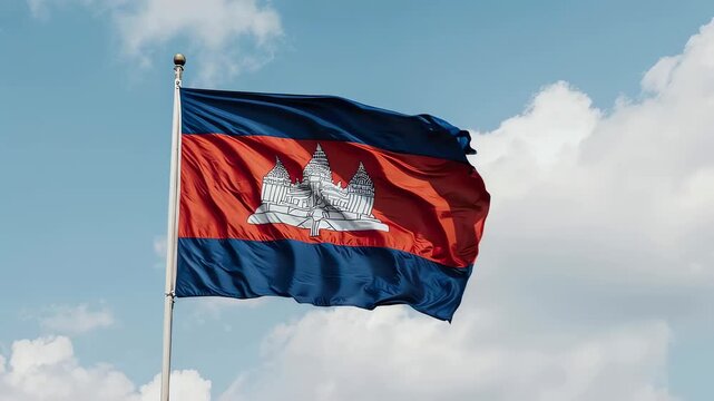 The Cambodian flag, featuring the iconic Angkor Wat temple silhouette, waves proudly against a clear blue sky with scattered white clouds, symbolizing national pride and heritage