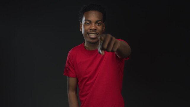 Friendly smiling young african american man pointing finger in studio against black wall wearing red shirt; confidence.