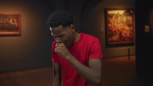 Young african american man in red shirt with hand on cheek for toothache in art gallery building; discomfort.