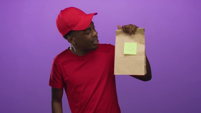Young man wearing red cap and red t shirt holding paper bag with smiley note in purple studio; happiness.
