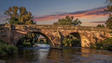 Obraz premium A serene stone bridge spans the tranquil river, its multiple arches reflected in the calm waters as golden hour casts a warm glow over the lush forest surroundings.