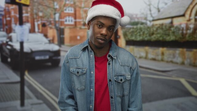 Man wearing red santa hat tilts head while gazing downward in denim jacket on city street; reflection calm.