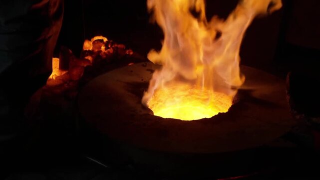 Close up of a worker skimming impurities from the surface of molten metal inside a burning furnace crucible