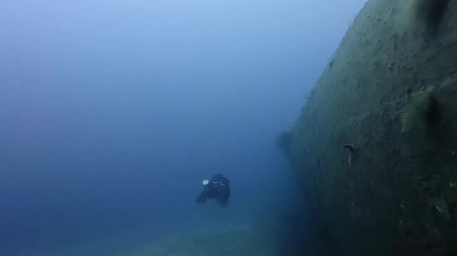 Submerged Scuba Diver Exploring Massive Rusting Steel Hull of Sunken Shipwreck Deep in Blue Winter Mediterranean Sea Waters Low Angle Perspective Capturing Atmospheric Mystery of Lost Maritime History