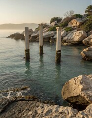 Fototapeta premium A serene rocky coastline features concrete pillars extending into the sea, protected by a breakwater structure amidst a tranquil morning atmosphere.