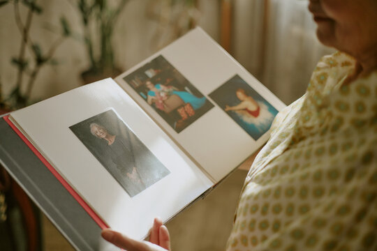 Senior Caucasian woman holding photo album, viewing old photographs with gentle expression, partial face visible, standing indoors near blurred background, reminiscing memories