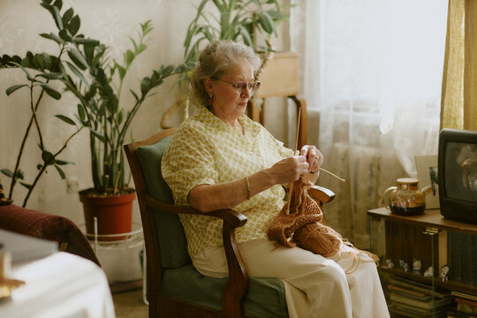 Senior Caucasian woman sitting in armchair knitting with yarn, wearing eyeglasses and earrings, concentrating on craft project in living room with houseplants and television visible