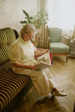 Senior Caucasian woman sitting on sofa looking through photo album, wearing glasses and focusing on old photographs, wooden floor and houseplants visible in background