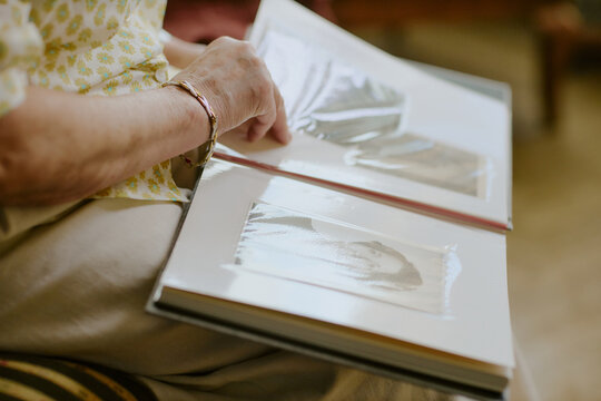Senior Caucasian woman sitting and browsing photo album, gently turning page with left hand, focusing on black and photographs, capturing nostalgic family memories