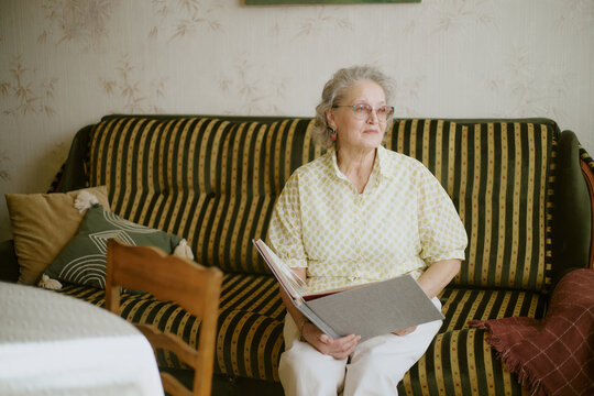 Senior Caucasian woman sitting on striped sofa holding large photo album, gazing thoughtfully to side, wearing eyeglasses, short gray hair, appearing relaxed in home setting