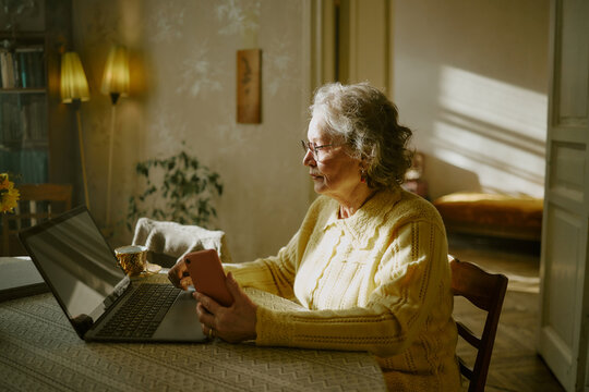 Senior Caucasian woman sitting at table using laptop and holding smartphone, wearing glasses and looking at computer screen, sunlight streaming through window in home interior