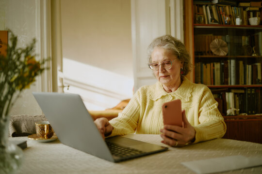 Senior Caucasian woman sitting at table using laptop and holding smartphone, wearing eyeglasses and concentrating on screen, bookshelf and cup visible in background