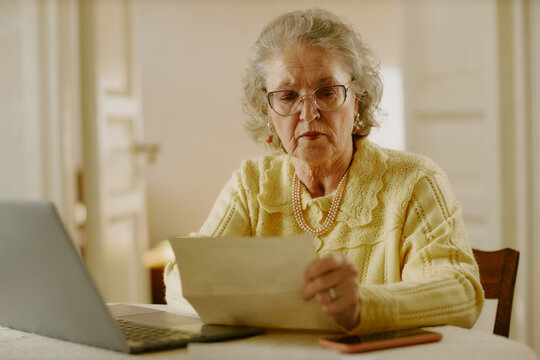 Senior Caucasian woman reading document while sitting at table with open laptop and smartphone, wearing glasses and jewelry, appearing focused and engaged in paperwork at home