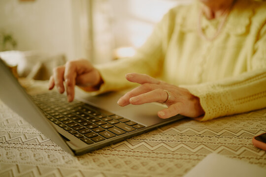 Senior Caucasian woman typing on laptop at table, hands visible with wedding ring, focusing on keyboard, demonstrating technology use by older adult, partial body shot