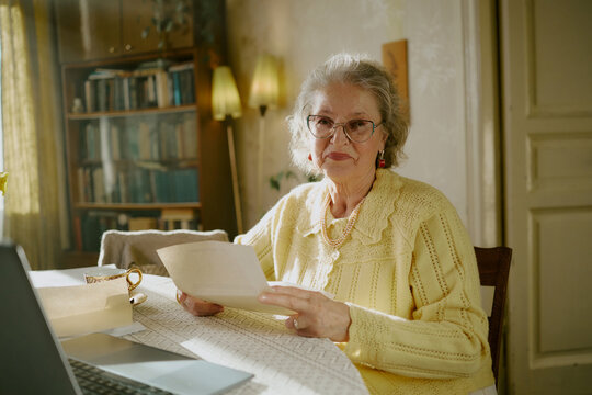 Portrait of senior Caucasian woman wearing eyeglasses sitting at table holding paper document and looking into camera with gentle smile, laptop and cup visible on table