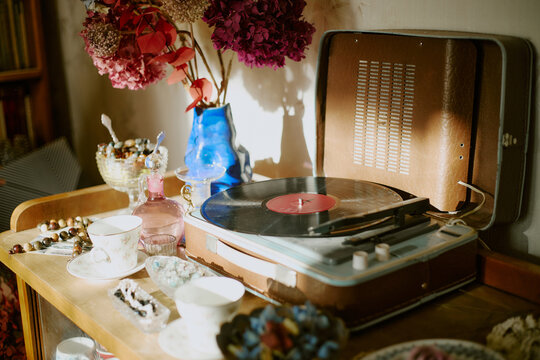 Vintage record player spinning vinyl on wooden table surrounded by porcelain teacups, glass vases with dried flowers, candy dishes and decorative objects in cozy indoor setting