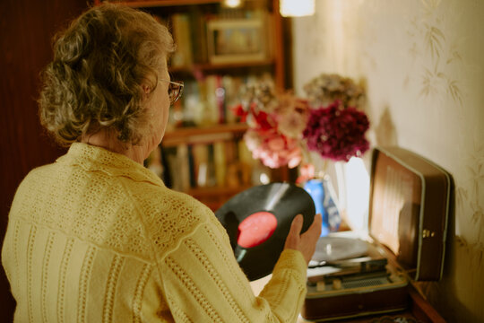 Senior Caucasian woman standing indoors holding vinyl record preparing to place it on vintage turntable with bookshelf and vase of flowers visible in background