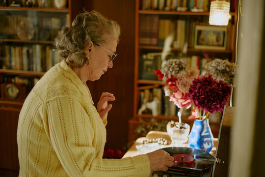 Senior Caucasian woman listening to vinyl record, standing beside turntable in cozy home library, touching record with hand, surrounded by bookshelves and decorative flowers