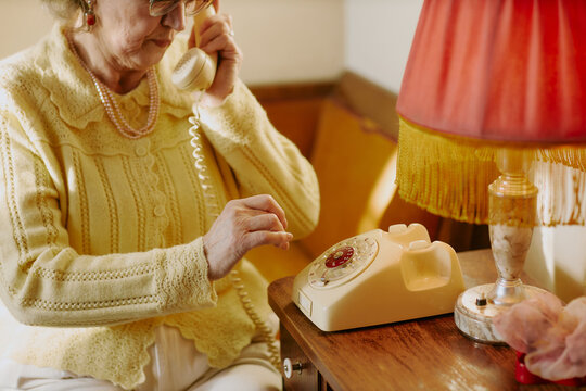 Senior Caucasian woman sitting indoors using rotary phone, holding receiver to ear while dialing number with finger, wearing eyeglasses and jewelry, vintage style setting visible
