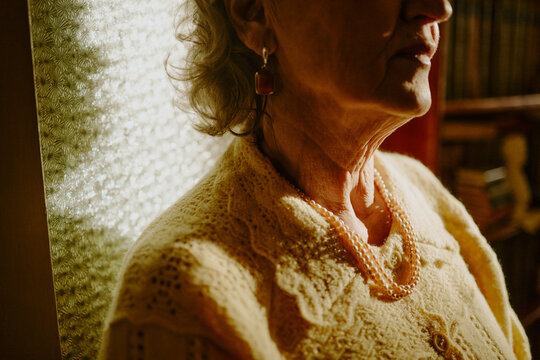 Senior Caucasian woman sitting indoors, wearing earrings and pearl necklace, partially facing camera with soft light highlighting facial features and textured background
