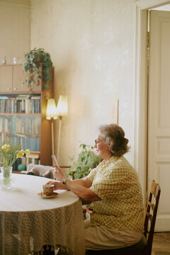 Senior Caucasian woman sitting at table using phone, smiling and interacting with device, cup of tea and vase with flowers on table, bookshelf and plants in background