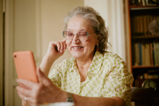 Portrait of senior Caucasian woman smiling while holding smartphone, sitting indoors, wearing eyeglasses, engaging in video call or chatting, showing joyful facial expression